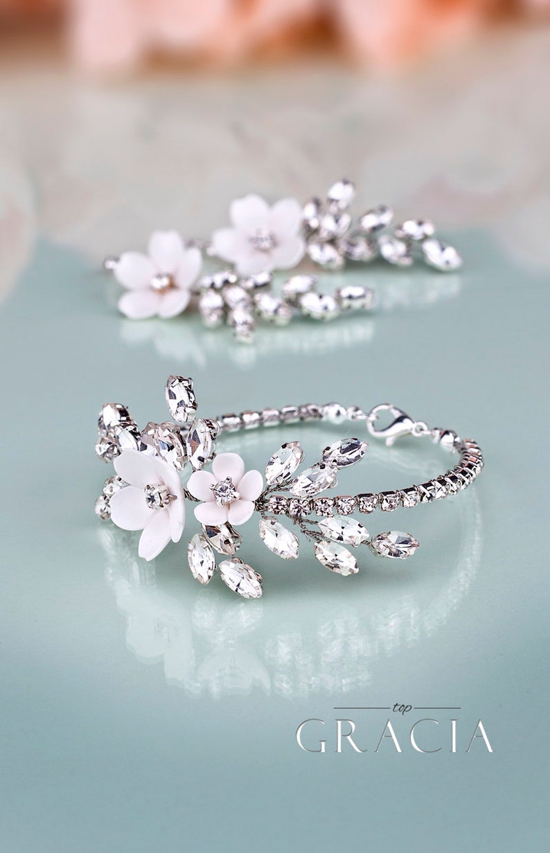K&ouml;nnte beinhalten: Ein silbernes Armband und ein passendes Haaraccessoire, beide mit wei&szlig;en Blumenakzenten und funkelnden Kristallen verziert. Das Armband hat ein filigranes Kettendesign, ideal f&uuml;r Hochzeiten. Das Wort "GRACIA" ist sichtbar.