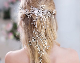 May include: A silver-toned bridal hairpiece with a floral and leaf design. The headpiece features a central floral cluster with cascading strands of crystals and smaller floral accents, creating an elegant, flowing look. The hair accessory is designed to be worn at the back of the head.