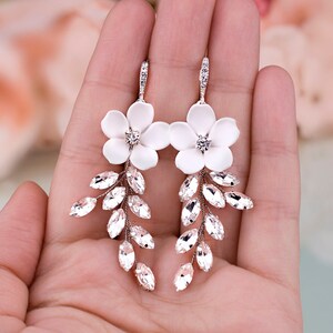 K&ouml;nnte beinhalten: Elegante wei&szlig;e Blumenohrringe mit Kristallakzenten. Jeder Ohrring hat eine wei&szlig;e Blume mit einem zentralen Kristall und Kristallbl&auml;ttern. Die Ohrringe werden in einer Hand gehalten, was ihr zartes Design zeigt.