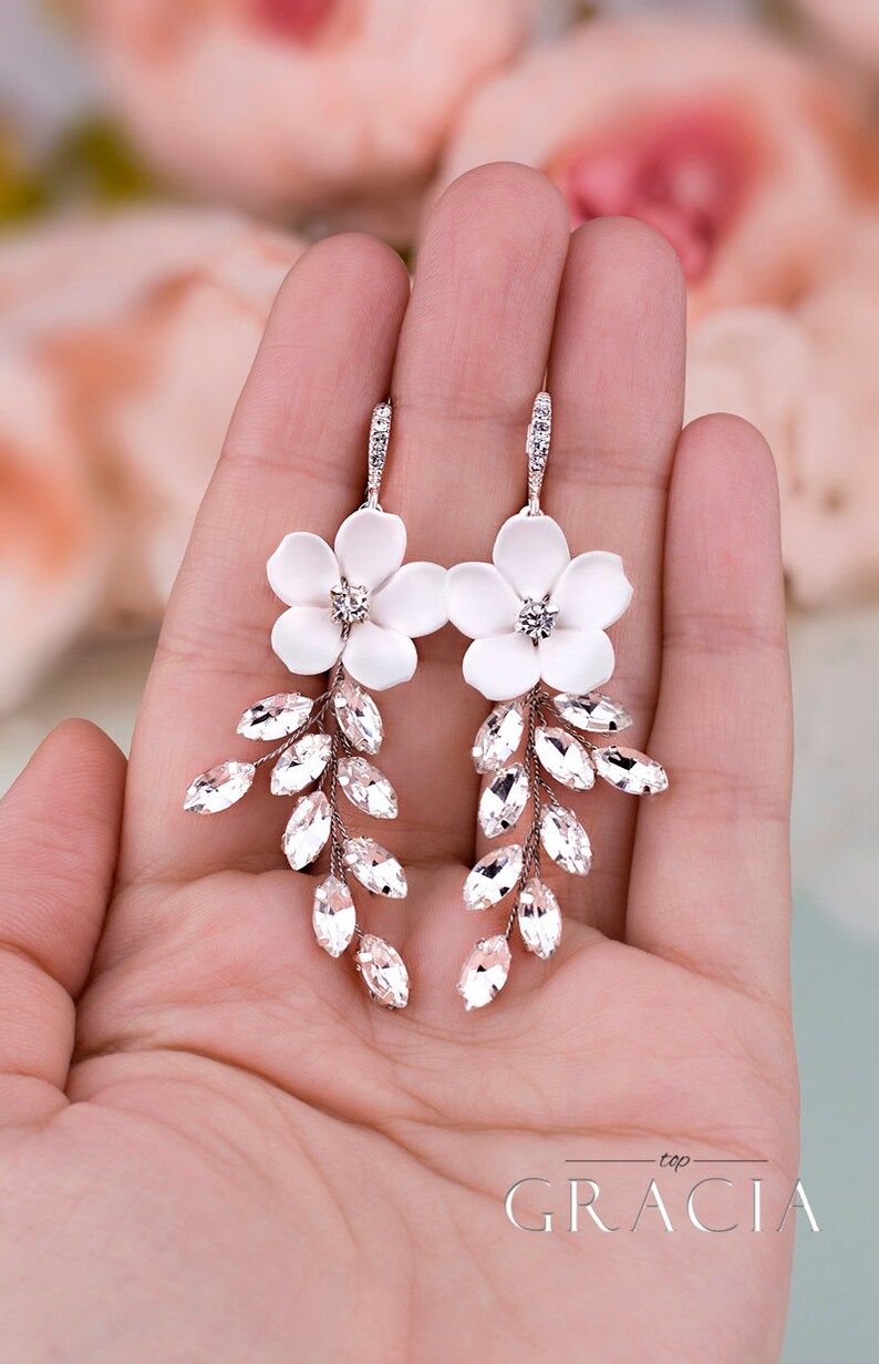 K&ouml;nnte beinhalten: Elegante wei&szlig;e Blumenohrringe mit Kristallakzenten. Jeder Ohrring hat eine wei&szlig;e Blume mit einem zentralen Kristall und Kristallbl&auml;ttern. Die Ohrringe werden in einer Hand gehalten, was ihr zartes Design zeigt.