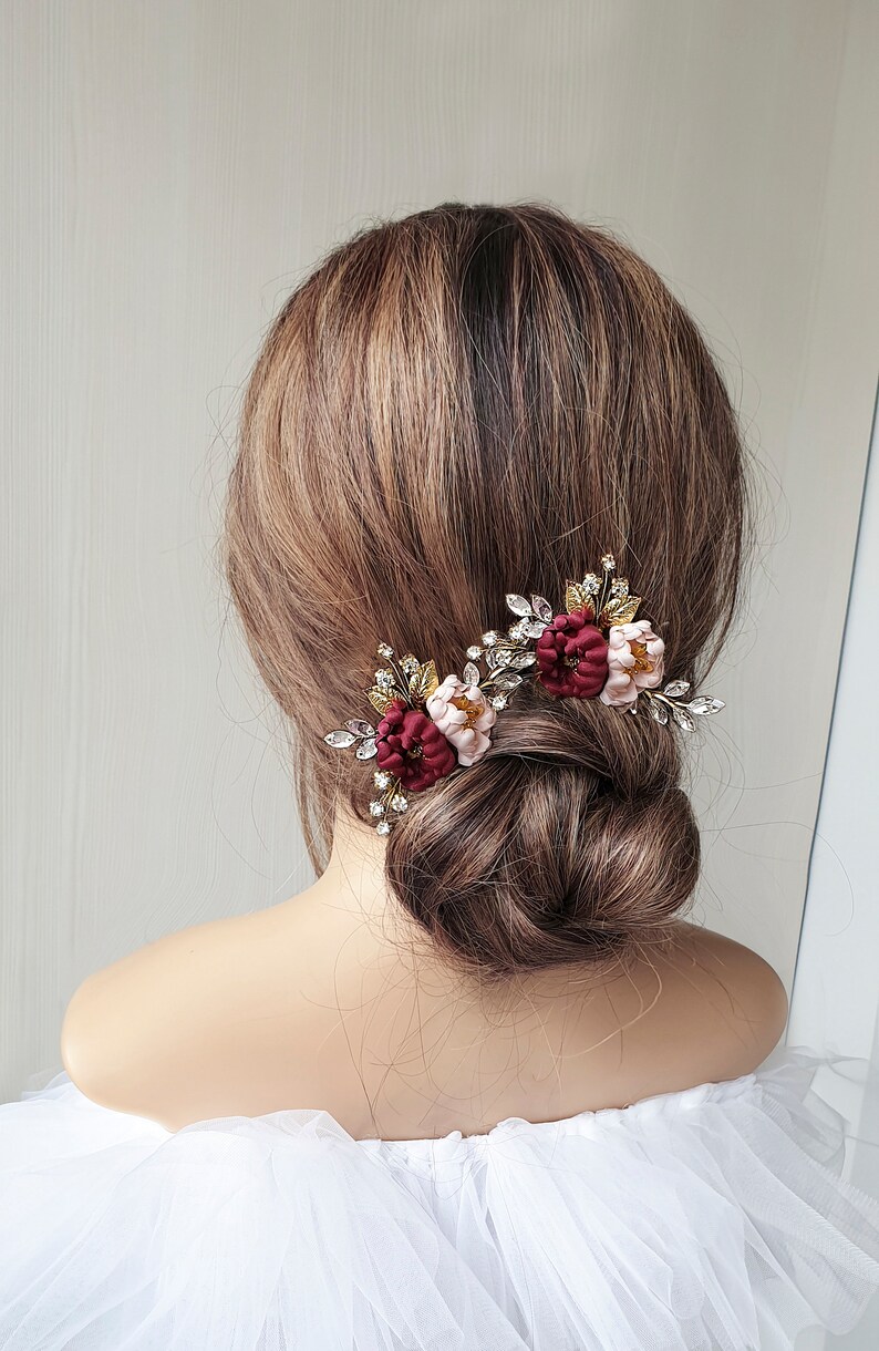 May include: Two burgundy and blush floral hair clips with gold and crystal embellishments. The clips are worn in a woman's hair.