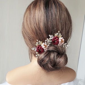 May include: Two burgundy and blush floral hair clips with gold and crystal embellishments. The clips are worn in a woman's hair.