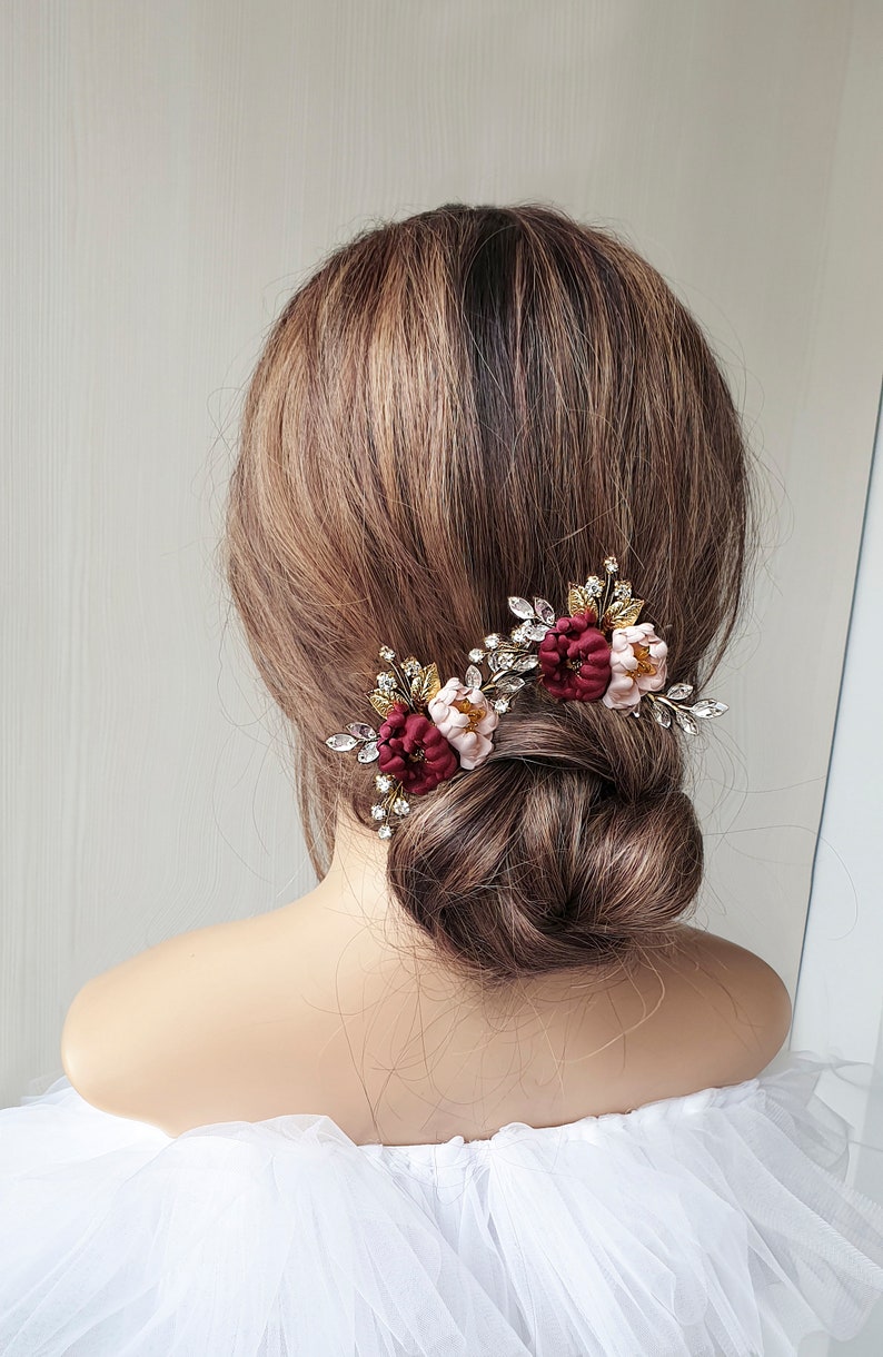 May include: Two burgundy and blush floral hair clips with gold and crystal embellishments. The clips are worn in a woman's hair.