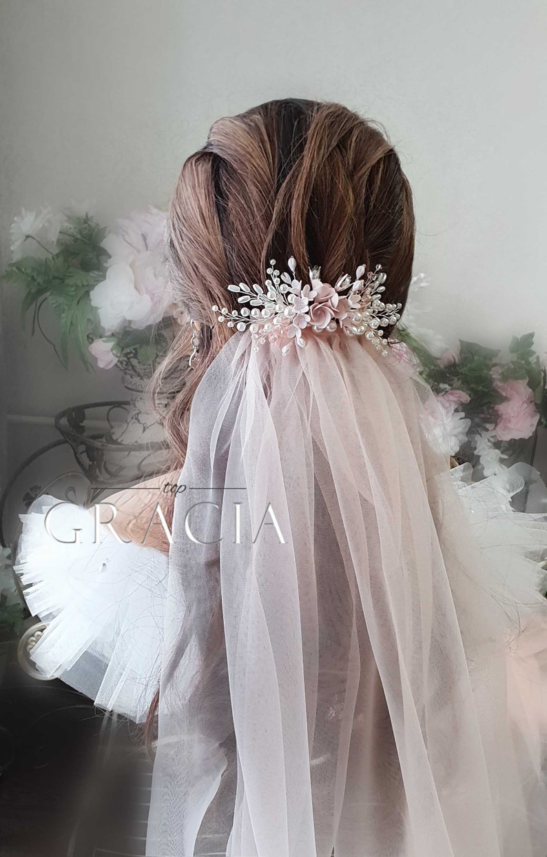 May include: A delicate bridal veil with a floral hair comb featuring pink flowers and pearl accents. The veil is made of soft tulle and is perfect for a wedding or special occasion.
