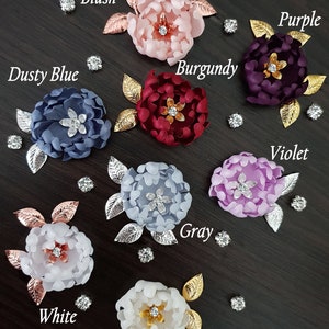 May include: Assortment of ten fabric flower hair clips in various colors, including blush, burgundy, dusty blue, gray, ivory, purple, violet, and white. Each flower has a rhinestone center and gold or silver leaf accents.