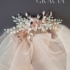 May include: A blush pink bridal veil with a delicate floral comb featuring blush pink and white flowers, pearls, and rhinestones. The comb is attached to the veil with a silver wire.