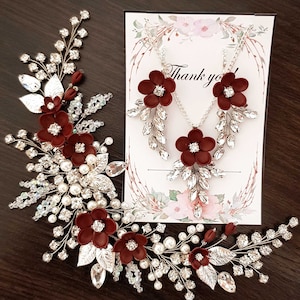 K&ouml;nnte beinhalten: Ein elegantes Schmuckset mit floralem Design. Das Set beinhaltet eine Halskette und Ohrringe mit burgunderfarbenen Blumenakzenten, klaren Kristallen und silberfarbenem Metall. Ein passendes Haarteil mit Perlen und Kristallen ist ebenfalls vorhanden, mit einer Karte mit "Danke".