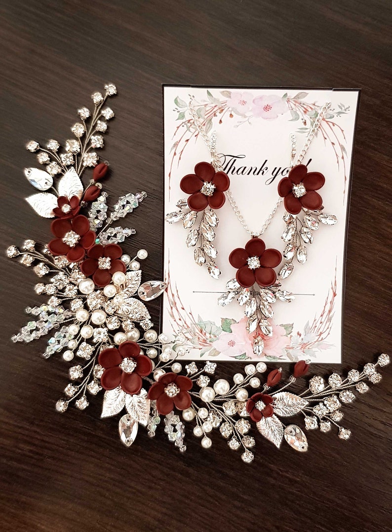 K&ouml;nnte beinhalten: Ein elegantes Schmuckset mit floralem Design. Das Set beinhaltet eine Halskette und Ohrringe mit burgunderfarbenen Blumenakzenten, klaren Kristallen und silberfarbenem Metall. Ein passendes Haarteil mit Perlen und Kristallen ist ebenfalls vorhanden, mit einer Karte mit "Danke".