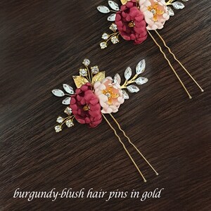 May include: Two gold hair pins with burgundy and blush fabric flowers and rhinestones. The pins are on a dark brown wooden surface.