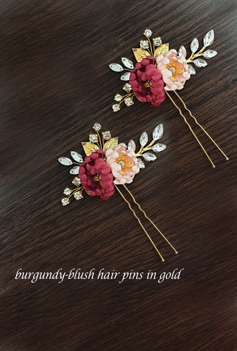 May include: Two gold hair pins with burgundy and blush fabric flowers and rhinestones. The pins are on a dark brown wooden surface.