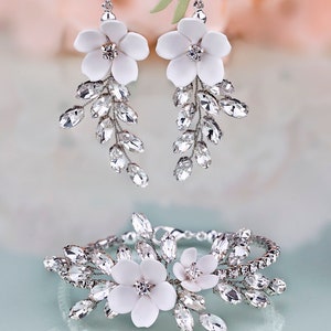 K&ouml;nnte beinhalten: Elegantes Brautschmuck-Set mit wei&szlig;en Blumenohrringen und einem passenden Armband. Die Ohrringe und das Armband sind mit funkelnden, klaren Kristallen und zarten wei&szlig;en Blumen verziert, was einen raffinierten Look erzeugt.