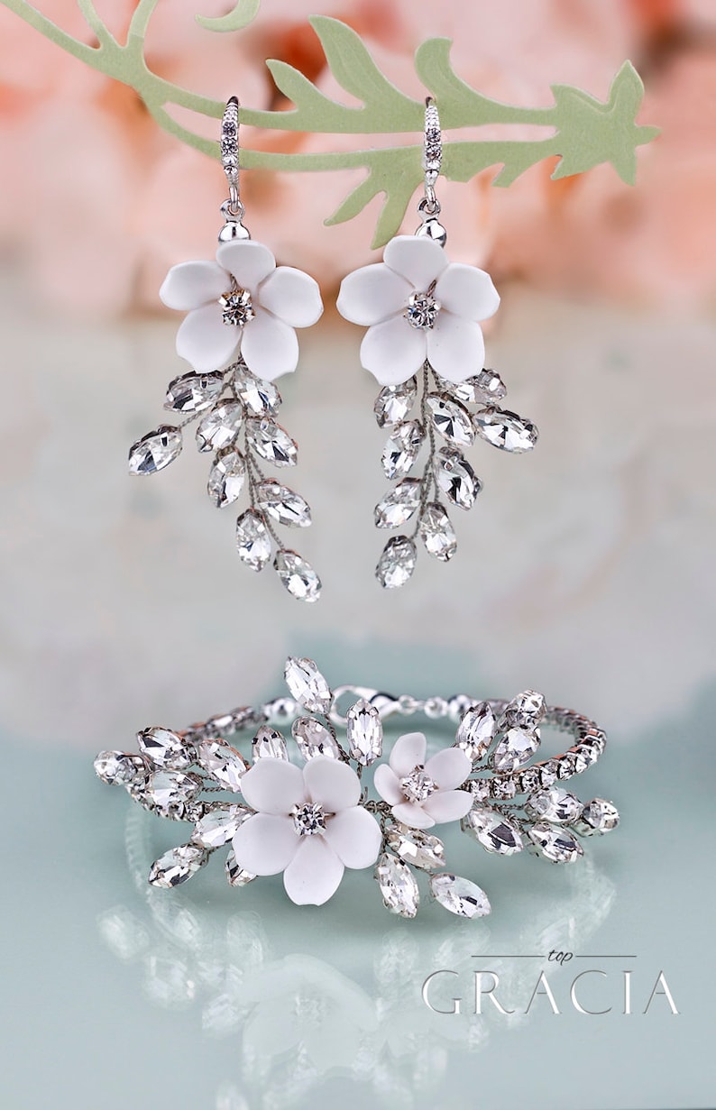 K&ouml;nnte beinhalten: Elegantes Brautschmuck-Set mit wei&szlig;en Blumenohrringen und einem passenden Armband. Die Ohrringe und das Armband sind mit funkelnden, klaren Kristallen und zarten wei&szlig;en Blumen verziert, was einen raffinierten Look erzeugt.