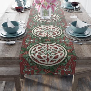 May include: A red and green table runner with a repeating Celtic knot design. The runner features a central pattern of cream-colored knots surrounded by green leaves and red berries. The table is set with blue bowls, white plates, and wine glasses.