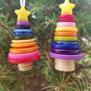 Puede incluir: Dos adornos de árbol de Navidad de botones coloridos. Cada árbol está hecho de una pila de botones coloridos con una estrella amarilla en la parte superior. Los árboles están colgados de una cinta blanca.