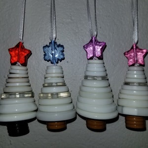 Puede incluir: Cuatro adornos de árbol de Navidad de botones blancos con diferentes colores de estrella y copo de nieve. Los adornos son rojo, azul, rosa y morado.
