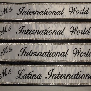 May include: Four white satin sashes with black trim and silver rhinestones. The sashes are embroidered with the text "Ms. International World" and "Ms. Latina International".