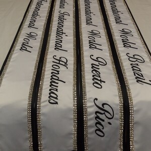 May include: Four white sashes with black trim and silver rhinestones. The sashes are labeled "Ms. International World Brazil", "Ms. International World Puerto Rico", "Ms. Latina International Honduras", and "Ms. International World".