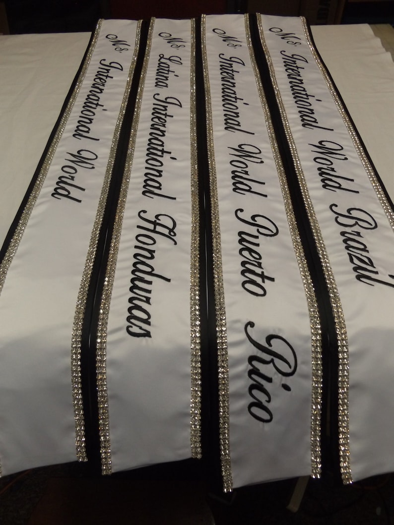 May include: Four white sashes with black trim and silver rhinestones. The sashes are labeled "Ms. International World Brazil", "Ms. International World Puerto Rico", "Ms. Latina International Honduras", and "Ms. International World".