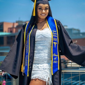 May include: A woman wearing a black graduation gown and a blue and gold sash with the text "Registered Nurse" and "Class of 2021".