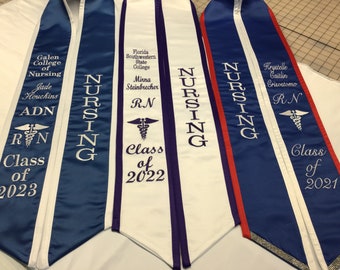 May include: Three white graduation sashes with blue and purple trim. The sashes are embroidered with the word "NURSING" and the names of the graduates. The sashes also include the graduate's degree, the name of the college, and the year of graduation.