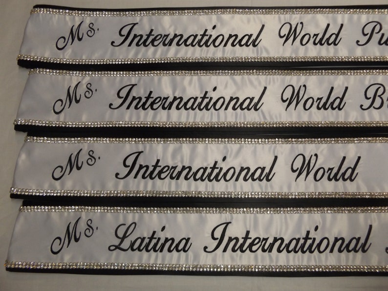 May include: Four white satin sashes with black trim and silver rhinestones. The sashes are embroidered with the text "Ms. International World" and "Ms. Latina International".