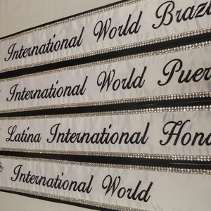 May include: Four white satin sashes with black trim and rhinestones. The sashes are labeled "Ms. International World" with the country name following: Brazil, Puerto, Honduras, and World.