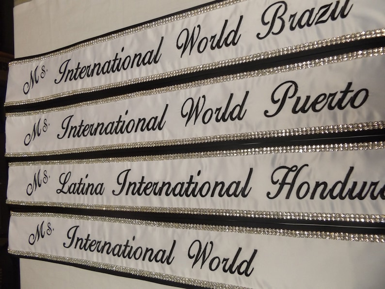 May include: Four white satin sashes with black trim and rhinestones. The sashes are labeled "Ms. International World" with the country name following: Brazil, Puerto, Honduras, and World.