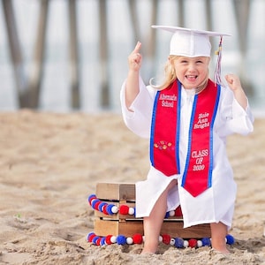May include: A young graduate wearing a white graduation gown and cap, a red and blue graduation sash with the text "Albemarle School", "Zoie Ann Bright", and "Class of 2020" embroidered on it. The graduate is sitting on a wooden crate with red, white, and blue pom-poms around it.