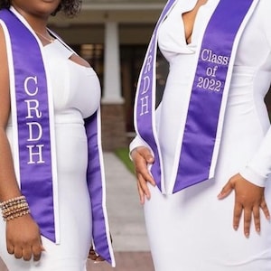 May include: Two people wearing white dresses and purple graduation sashes with white trim. The sashes have white text on them. One sash says "CRDH" and the other says "Class of 2022".