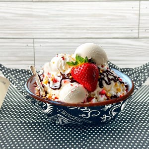 May include: A designer bowl cozy with a bowl of ice cream sundae. The sundae has vanilla ice cream, a large strawberry, chocolate sauce, and whipped cream. The bowl is navy blue with a white pattern and has handles.