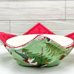 May include: A fabric bowl cozy with a Christmas tree and Santa Claus pattern on a green background. The rim and points are red with white polka dots. The interior is white.