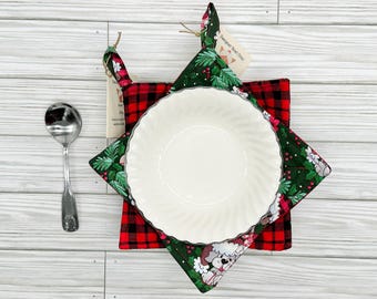 May include: A white ceramic bowl with a silver rim rests on a star-shaped pot holder. The pot holder features a red and black plaid pattern on one side and a green and white floral and puppy design on the other. A silver spoon is to the left.