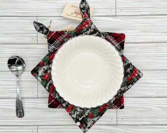 May include: A white ceramic bowl with a scalloped edge sits inside a plaid bowl cozy. The cozy is black, red, and gray with a Christmas theme. A silver spoon rests on the left side of the image.