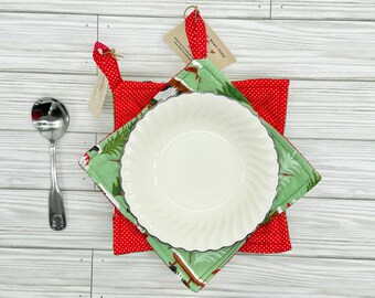 May include: A white ceramic bowl with a scalloped edge sits atop a green and red bowl cozy. The cozy has a red polka dot pattern on the bottom and a green Christmas tree design on top. A silver spoon rests to the side.