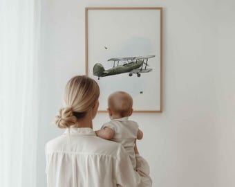 May include: A framed wall art print featuring a watercolor illustration of a vintage green biplane flying against a light blue sky background. The artwork is enclosed in a simple, light-colored wooden frame and is displayed on a plain white wall.
