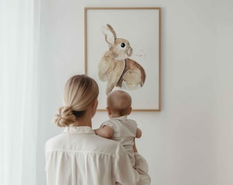 May include: A framed watercolor print of a rabbit in shades of brown and beige. The artwork is displayed on a white wall. A person is holding a baby in front of the artwork.