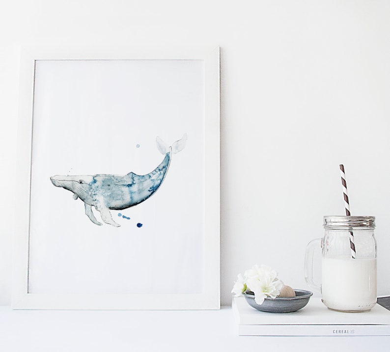 May include: A watercolor painting of a blue whale in a white frame. The whale is swimming to the right and has a light blue body with darker blue accents. The painting is on a white background.
