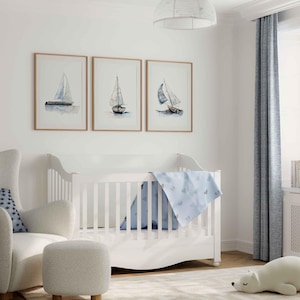 May include: A white crib with a blue and white patterned blanket. A white armchair with a blue and white patterned pillow sits next to the crib. Three framed watercolor paintings of sailboats hang on the wall behind the crib.