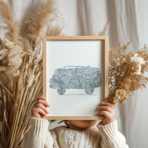 Op de afbeelding: Een ingelijste print met een lichtblauwe camperbus-silhouet gevuld met een wereldkaartontwerp. Het kunstwerk wordt vastgehouden door een persoon die een crèmekleurige trui draagt. Het frame is van lichtgekleurd hout.