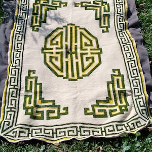 Op de afbeelding: Een crèmekleurige deken met een geometrisch ontwerp in groen en geel. De deken heeft een centraal cirkelvormig patroon en hoekontwerpen, allemaal omlijst door een rand van herhalende vierkanten. De deken ligt op een grijs oppervlak over gras.