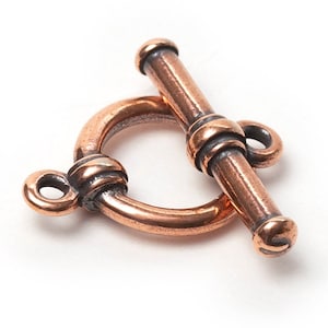 Può includere: Una chiusura a toggle color rame, un componente di gioielleria, è appoggiata su una superficie bianca. La chiusura presenta un anello circolare con una barra che scorre attraverso di esso. Il metallo ha un aspetto leggermente invecchiato.