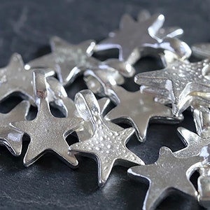 Könnte beinhalten: Eine Ansammlung silberfarbener Sternanhänger mit einer Schlaufe zum Aufhängen. Die Anhänger haben ein fünfzackiges Sterndesign und liegen auf einer dunkelgrauen Oberfläche. Die Charms sind aus Metall und haben ein glänzendes Finish.