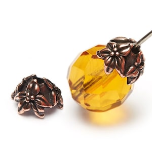 May include: Close-up of jewelry components. A faceted, amber-colored crystal bead is partially strung on a silver needle. A decorative, bronze-colored bead cap with a floral design sits next to the crystal bead. White background.