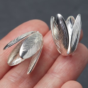 May include: Two silver-colored, tulip-shaped jewelry components. Each component has a feather-like design etched into the petals. The components are displayed against a dark background.