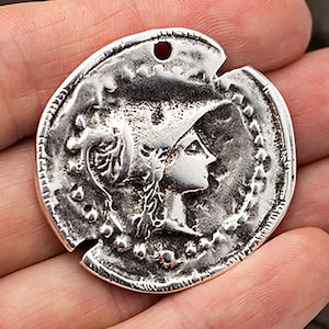 May include: A silver-colored coin pendant featuring a profile of a woman wearing a helmet. The coin has a distressed, antique finish with a small hole at the top for stringing. The coin's edge is textured with small dots.