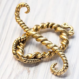May include: A gold-coloured decorative clasp with a twisted rope design. The clasp is in the shape of a figure eight, with a looped design on one end and a curled end on the other. The clasp is on a white wooden surface.