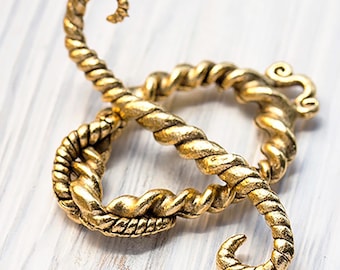 May include: A gold-colored, decorative clasp with a twisted rope design. The clasp is in the shape of a figure eight, with a looped design on one end and a curled end on the other. The clasp is on a white wooden surface.