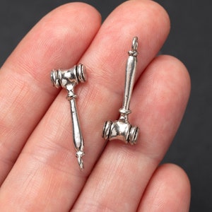 May include: Two small, silver-colored gavel charms. The gavels have a detailed design with a rounded head and a long, slender handle. The charms are shown against a neutral background.