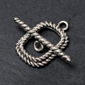 Op de afbeelding: Een zilverkleurige toggle sluiting met een touwachtig ontwerp. De sluiting heeft een cirkelvormige lus en een staaf die door een vierkante lus kruist. Het metaal heeft een verouderde, antieke uitstraling, tegen een donkere achtergrond.