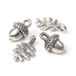 May include: Four silver-coloured metal charms are shown. Two are acorns, and two are oak leaves. The acorn charms have textured caps and smooth bodies. The leaf charms have detailed veining. Each charm has a loop for attaching to jewellery or other crafts.